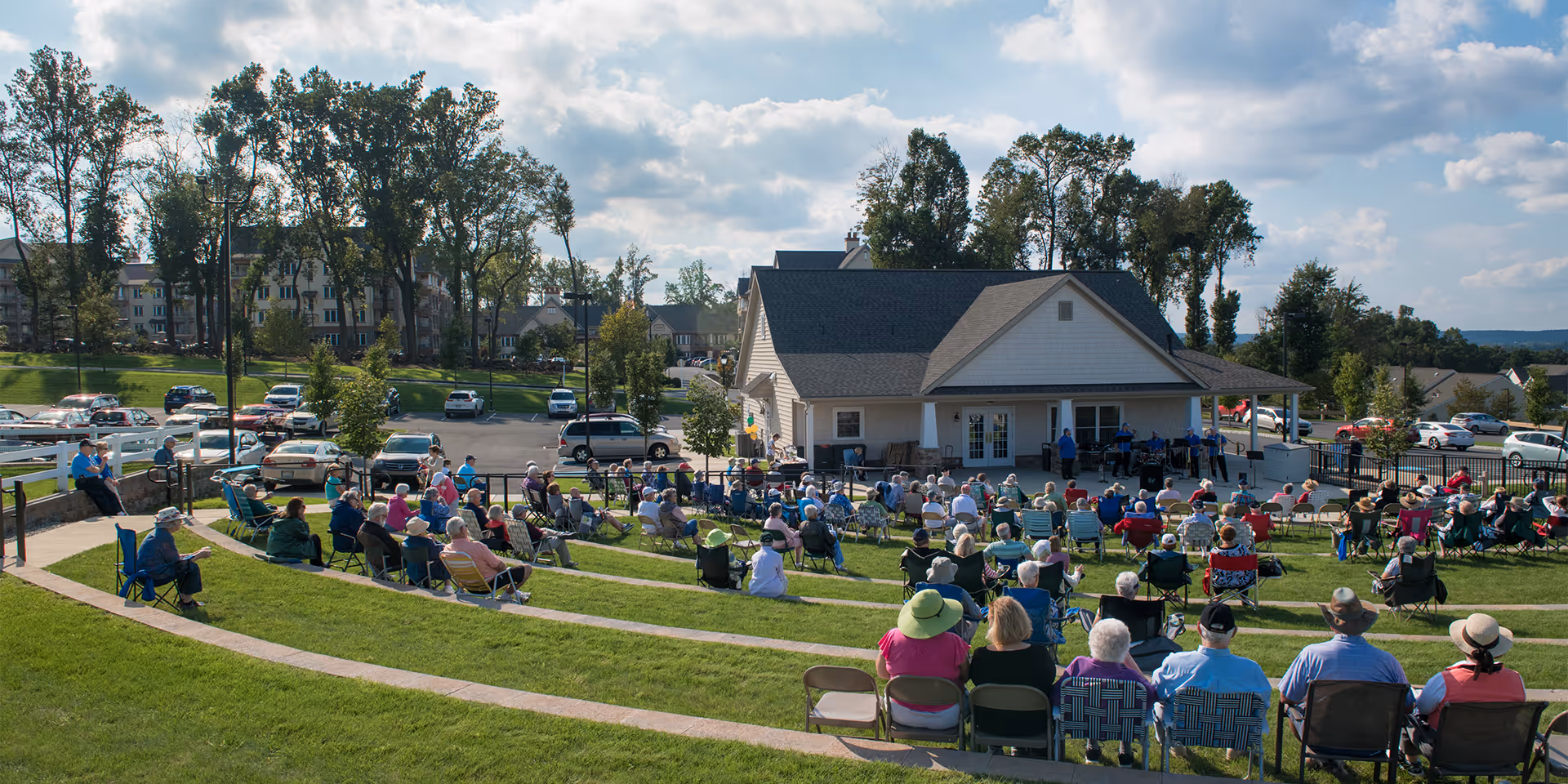 A crowd of older adults seated on tiered grassy lawn watching a performance in front of a community building.