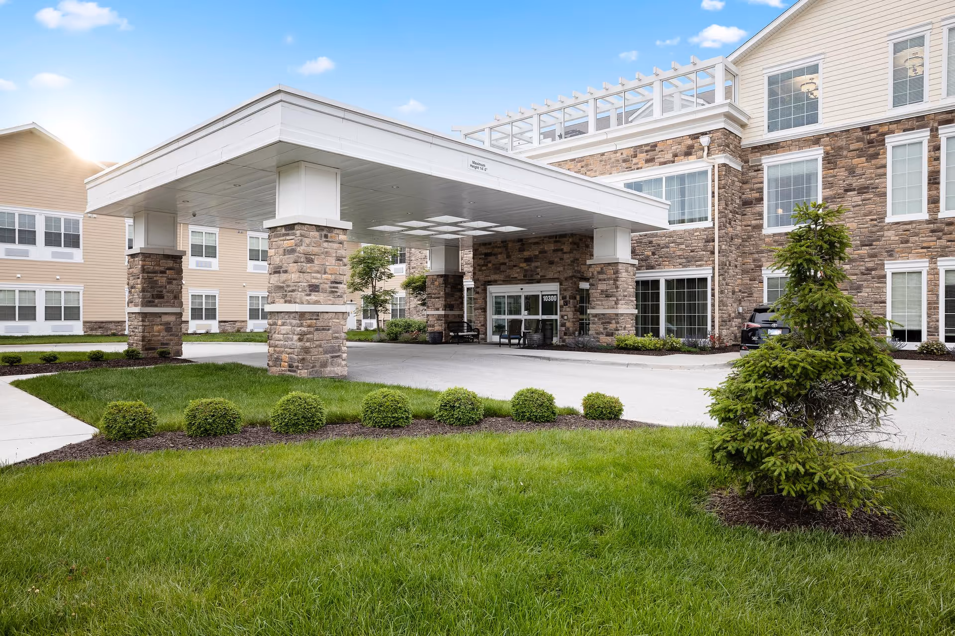 Exterior view of a senior living facility entrance with a covered drop-off area supported by stone pillars, surrounded by green grass, small bushes, and a small tree under a clear blue sky.