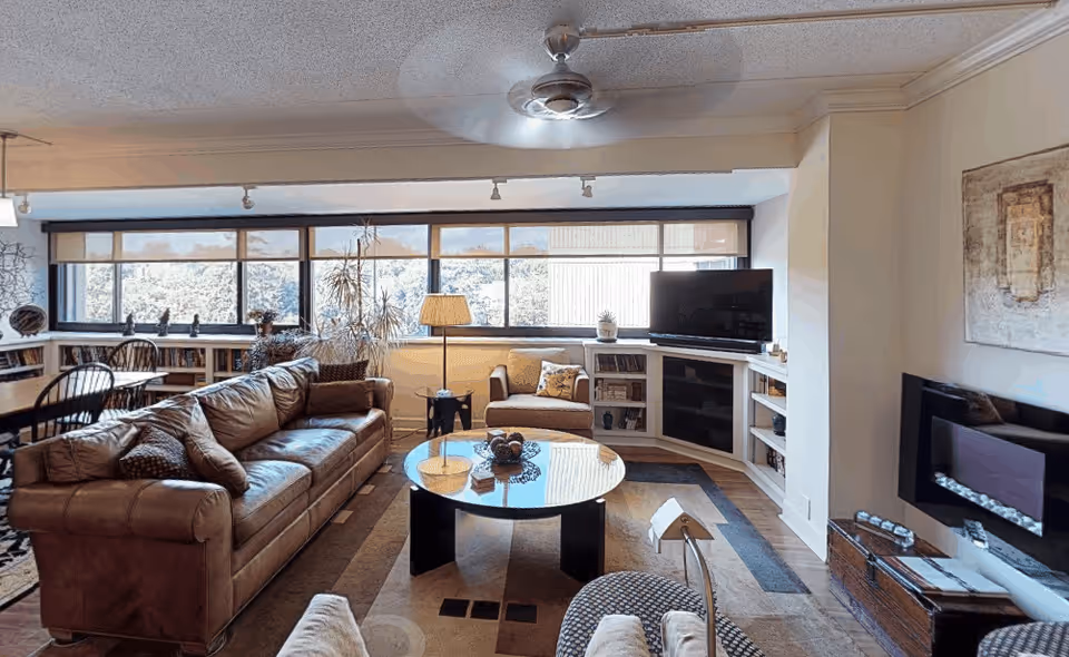 A cozy living room with a large brown leather sofa, an armchair, a round glass coffee table, and a TV on a corner stand. The room features large windows with a view of trees outside, a ceiling fan, a floor lamp, bookshelves, and a modern wall-mounted fireplace.
