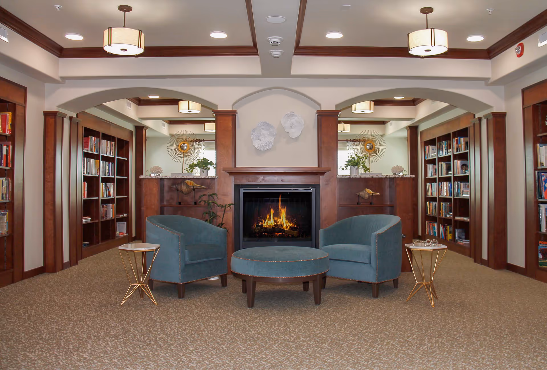 A cozy interior lounge area with two teal armchairs and a matching round ottoman arranged around a lit fireplace. The fireplace is framed with wood and has decorative white wall art above it. On either side of the fireplace are built-in wooden bookshelves filled with books. The room has beige carpet, recessed lighting, and ceiling-mounted light fixtures.