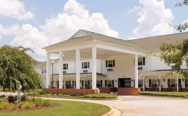 Front entrance of a two-story white senior living building with a large covered porte-cochère and landscaped driveway.