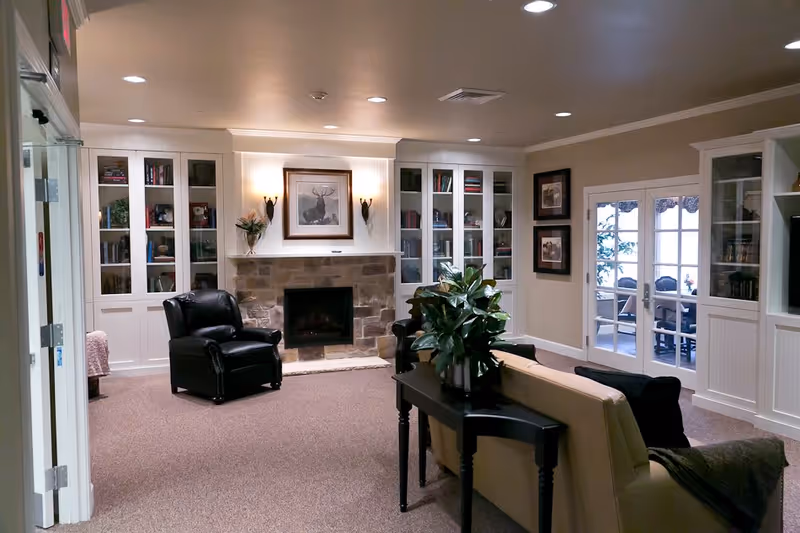 A cozy living room area in Meadowbrook Memory Care Community featuring a stone fireplace with a framed deer picture above it, flanked by built-in white bookshelves filled with books and decorative items. There are two black leather armchairs near the fireplace, a beige sofa with a black cushion and a throw blanket, and a black console table with a green plant on it. French doors lead to another room with a table and chairs visible through the glass. The room has beige walls and carpeted flooring.