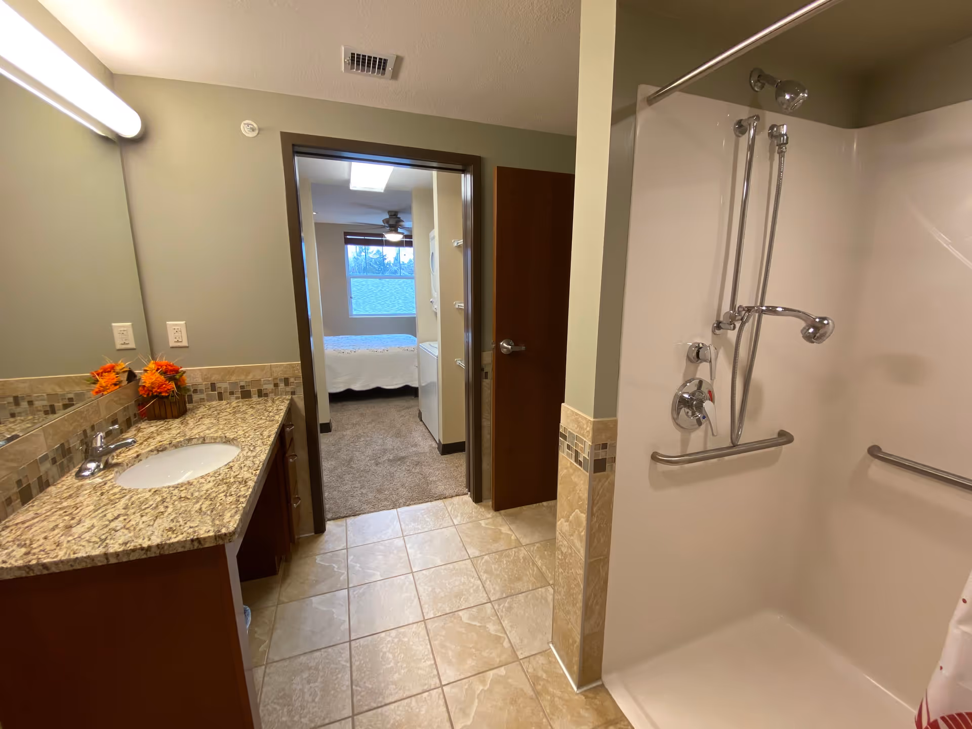 Bathroom with a granite countertop sink on the left and a walk-in shower with grab bars on the right. The bathroom floor is tiled, and there is a doorway leading to a bedroom with carpet and a window in the background.