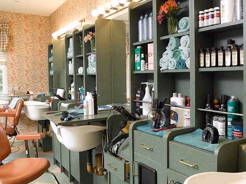 Interior view of a salon area with multiple green shelving units filled with hair care products, rolled towels, and styling tools. There are brown salon chairs and white sinks for washing hair, with large mirrors and bright lights above the counters.