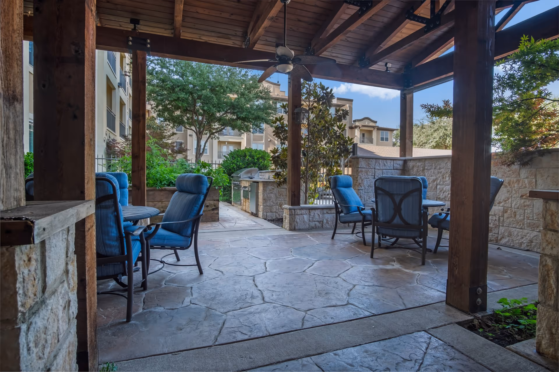 Covered outdoor patio area with stone flooring and wooden beams, furnished with blue cushioned chairs and round tables. In the background, there is a built-in stone grill and greenery including trees and bushes, with residential buildings visible beyond.