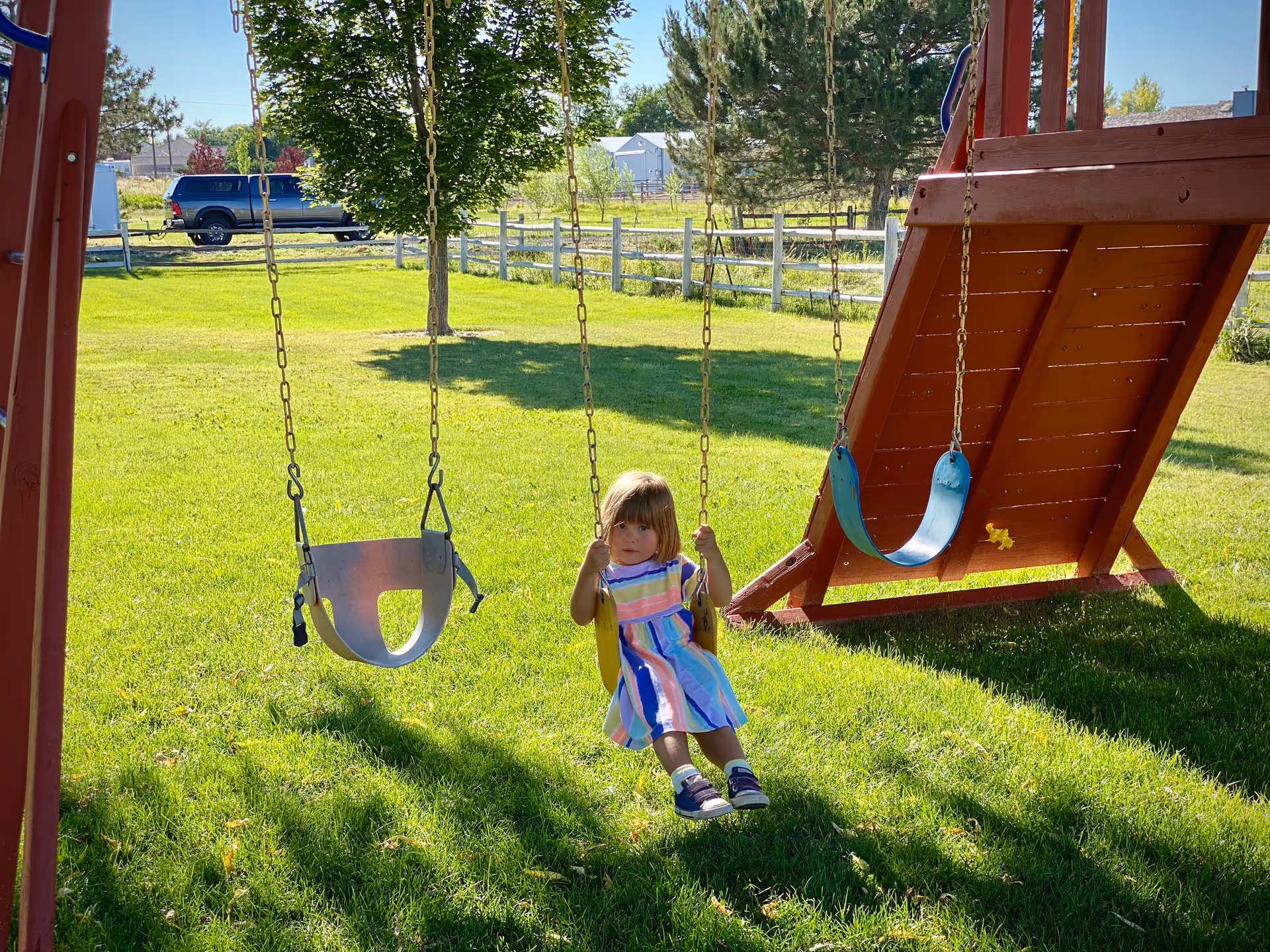 A child sits on a swing under a wooden playset on a sunny grassy yard.