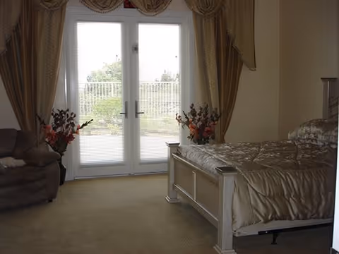 A bedroom with a large bed covered in a shiny beige comforter, a cushioned armchair to the left, and double glass doors with blinds leading outside. The room has beige walls and carpet, with floral arrangements on either side of the doors and decorative curtains above.