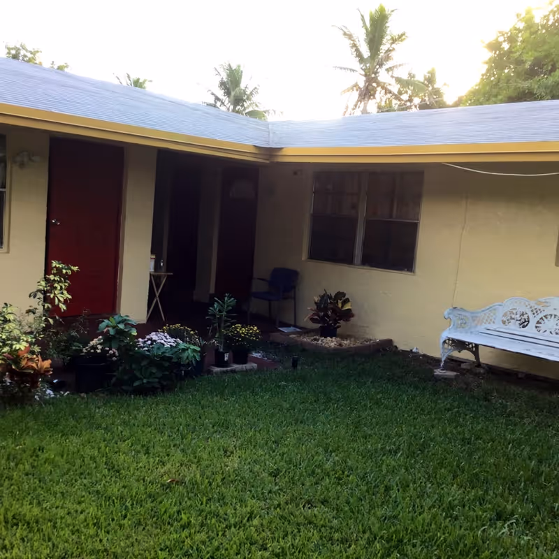 A small courtyard area with green grass, various potted plants, and a white decorative bench. The courtyard is surrounded by a single-story building with beige walls, red doors, and windows. There is a blue chair near one of the doors and some trees visible in the background.