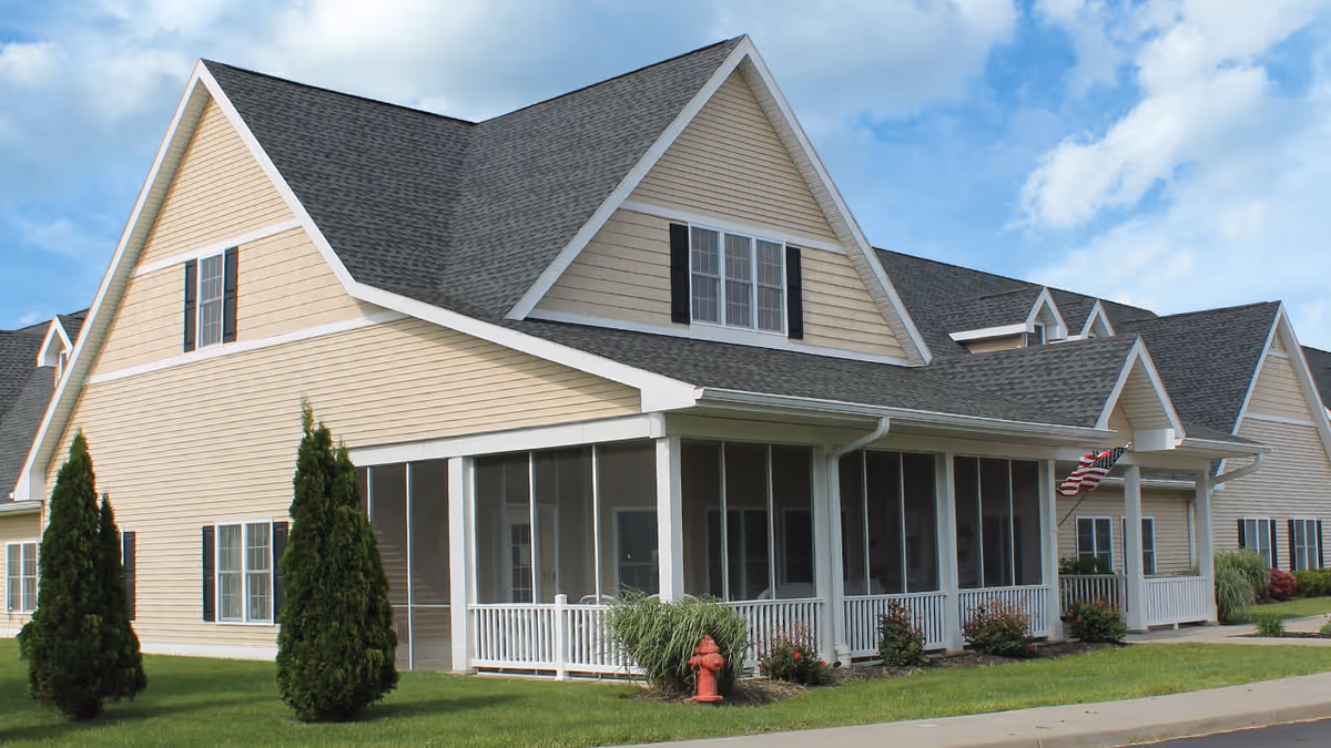 Exterior view of a beige two-story building with a gray shingled roof, white trim, and black window shutters. The building has a screened porch with white railings and an American flag hanging near the entrance. There are small bushes and two tall evergreen trees in the grassy area surrounding the building under a partly cloudy blue sky.