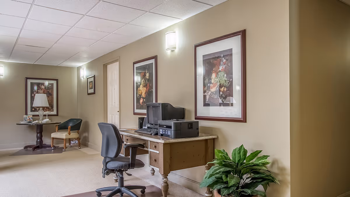 A hallway area in a senior living community with a wooden desk holding a computer, keyboard, and printer. There is a black office chair in front of the desk. The walls are beige with framed floral artwork and wall-mounted lights. In the background, there is a small round table with a lamp and a chair. A green potted plant is placed near the desk.