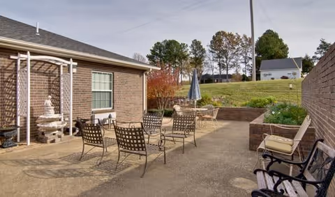 Outdoor brick courtyard patio with metal chairs and tables, a small fountain, umbrella, and surrounding brick walls and grassy slope with trees.