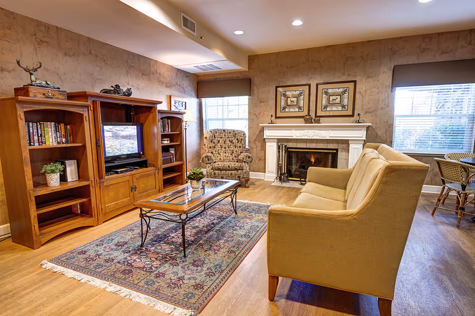 A cozy living room with a beige sofa, a patterned armchair, a glass coffee table on a decorative rug, a wooden entertainment center with a TV and books, and a white fireplace with two framed pictures above it. Two windows with blinds allow natural light into the room.