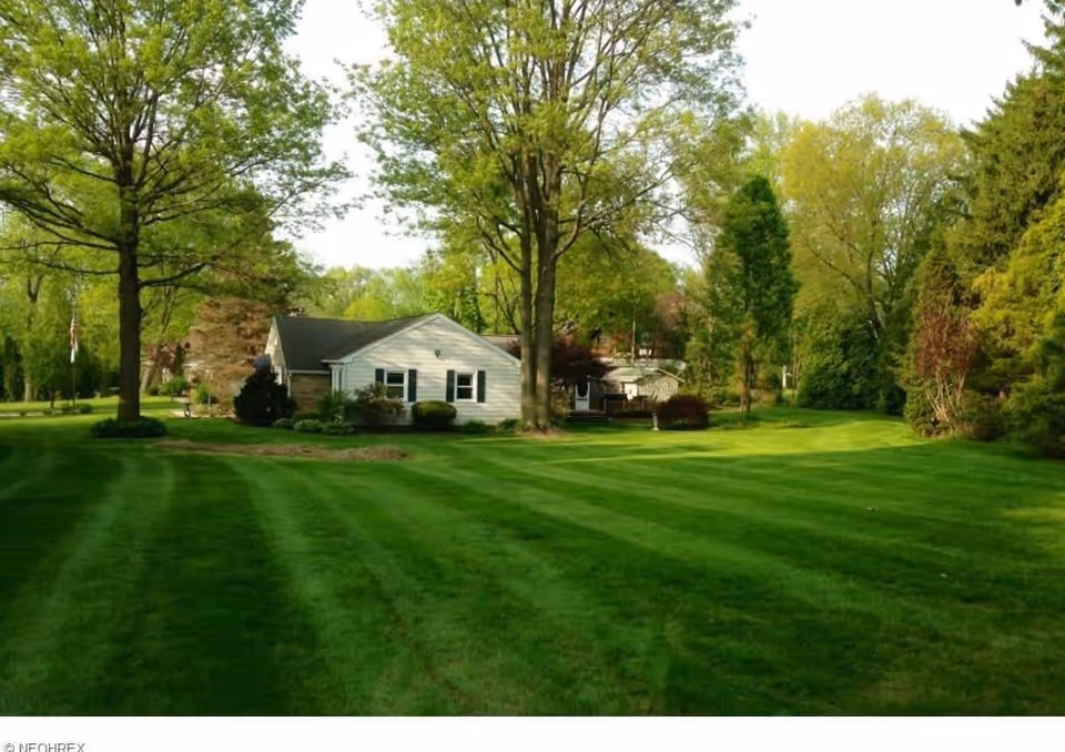A single-story white house surrounded by a large, well-maintained green lawn with striped mowing patterns. Several tall trees and shrubs are scattered around the property, and the scene is set on a bright, clear day.