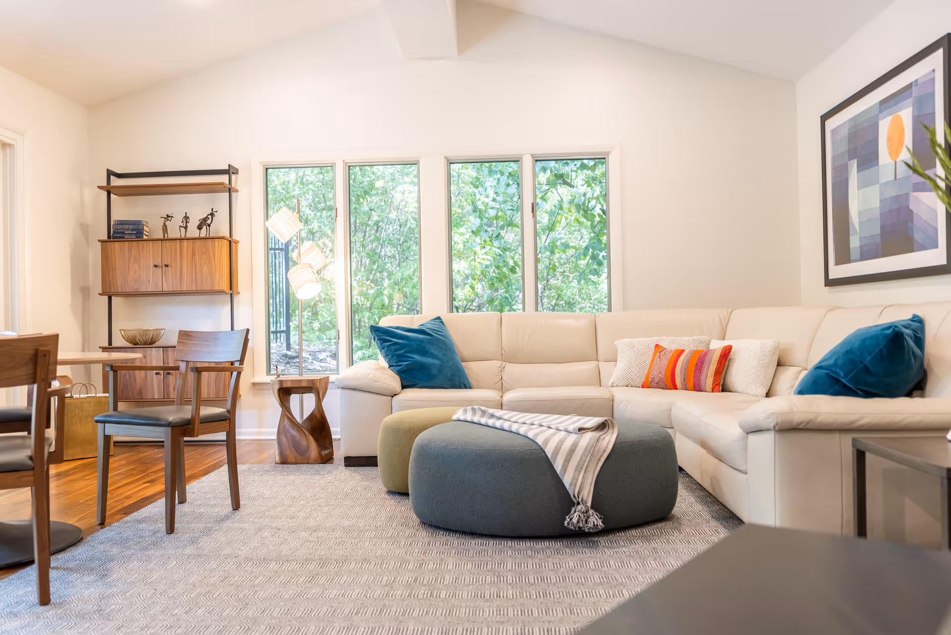 Bright living room with a large beige sectional sofa, colorful pillows, round ottomans, wooden chairs, shelving unit, and windows showing greenery outside.