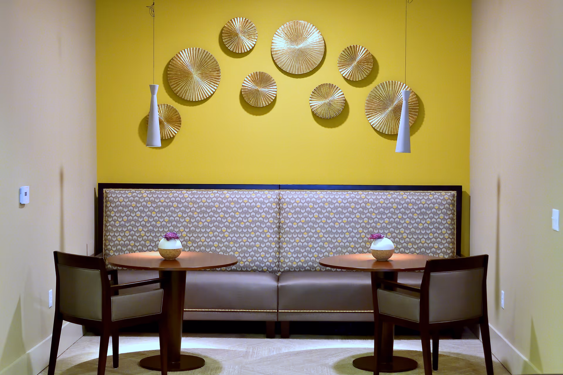 Small seating nook with a patterned bench, two round tables and chairs against a yellow wall decorated with gold circular wall art and pendant lights.
