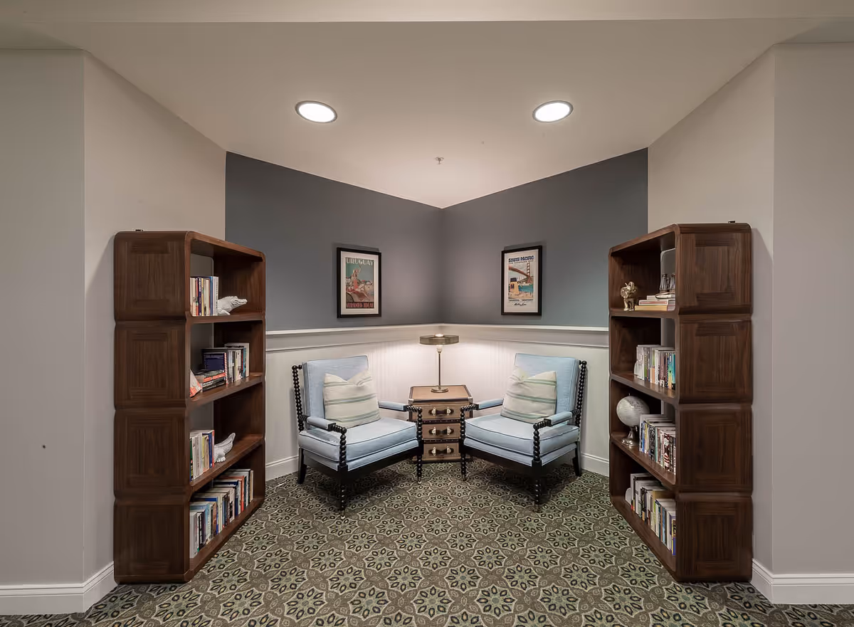 A cozy reading nook with two light blue armchairs facing each other, separated by a small wooden table with a lamp. The nook is framed by two wooden bookshelves filled with books on either side. The walls are painted gray and white with two framed vintage posters hanging above the chairs. The floor has a patterned carpet.