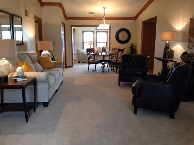 A spacious living room area with beige carpet and neutral-colored walls. The room features a light gray sofa with yellow and patterned throw pillows on the left, two black leather armchairs on the right, and a wooden side table with a lamp. In the background, there is a dining area with a round table and chairs, a large wall clock, and windows letting in natural light.