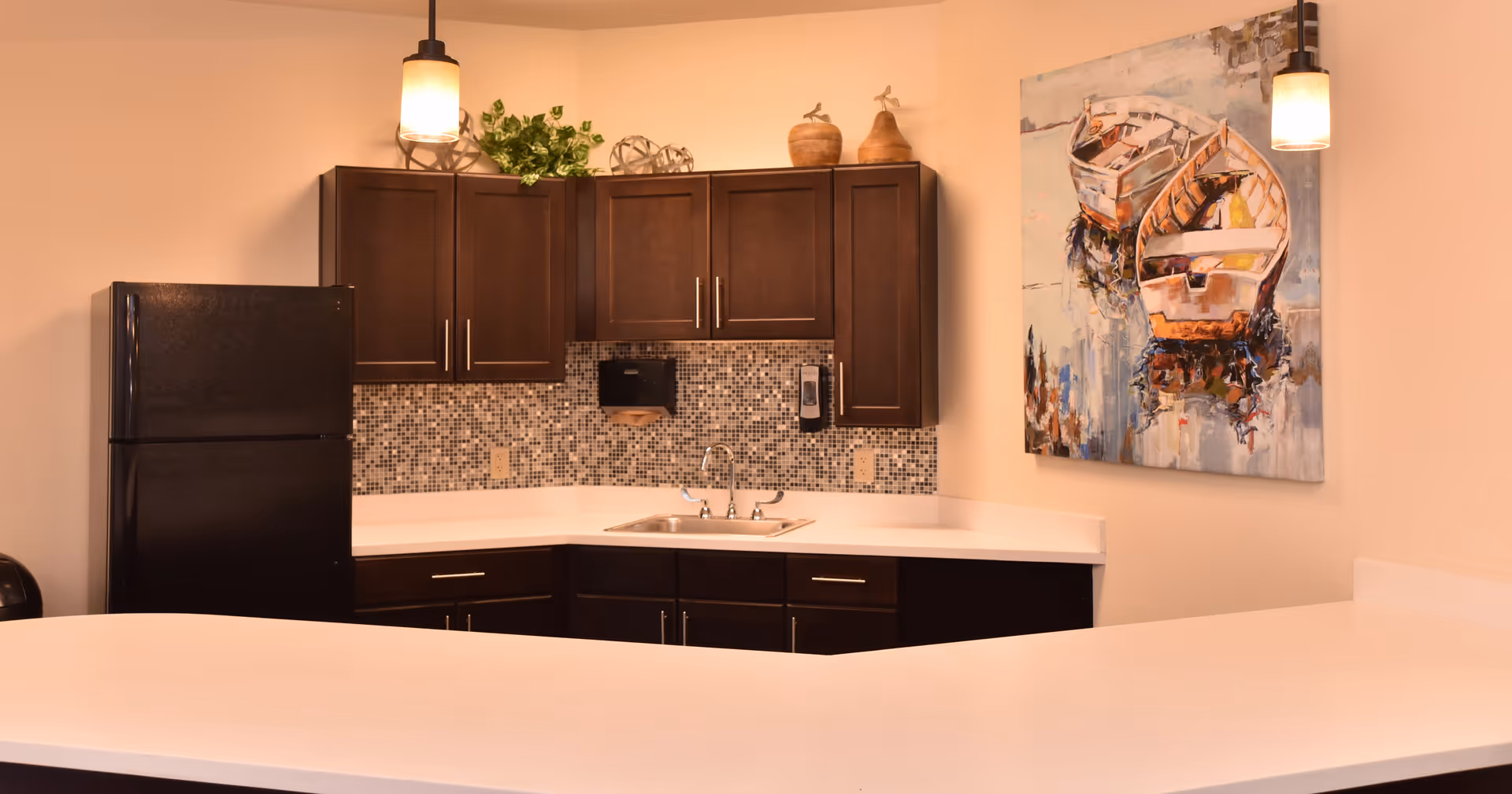 A modern kitchen area with dark wood cabinets, a black refrigerator, a mosaic tile backsplash, a stainless steel sink, and white countertops. There are two pendant lights hanging from the ceiling and a painting of boats on the wall.