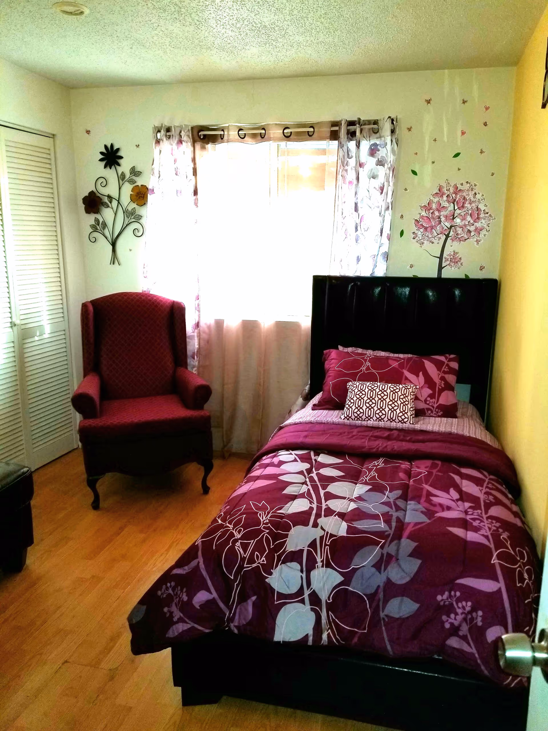 A small bedroom with a single bed covered in a purple floral comforter and matching pillows. Next to the bed is a red upholstered armchair. The room has wooden flooring, a window with floral curtains, and decorative wall art including a metal flower sculpture and a pink tree decal.