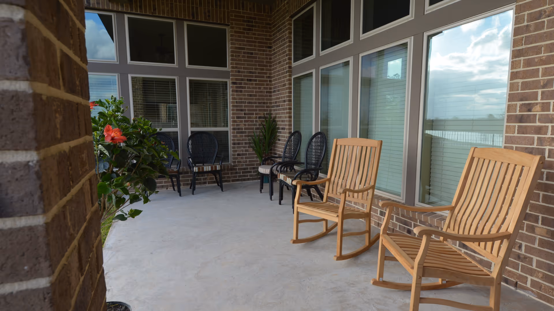 Covered brick patio with wooden rocking chairs, black wicker chairs, and potted plants along a row of windows.