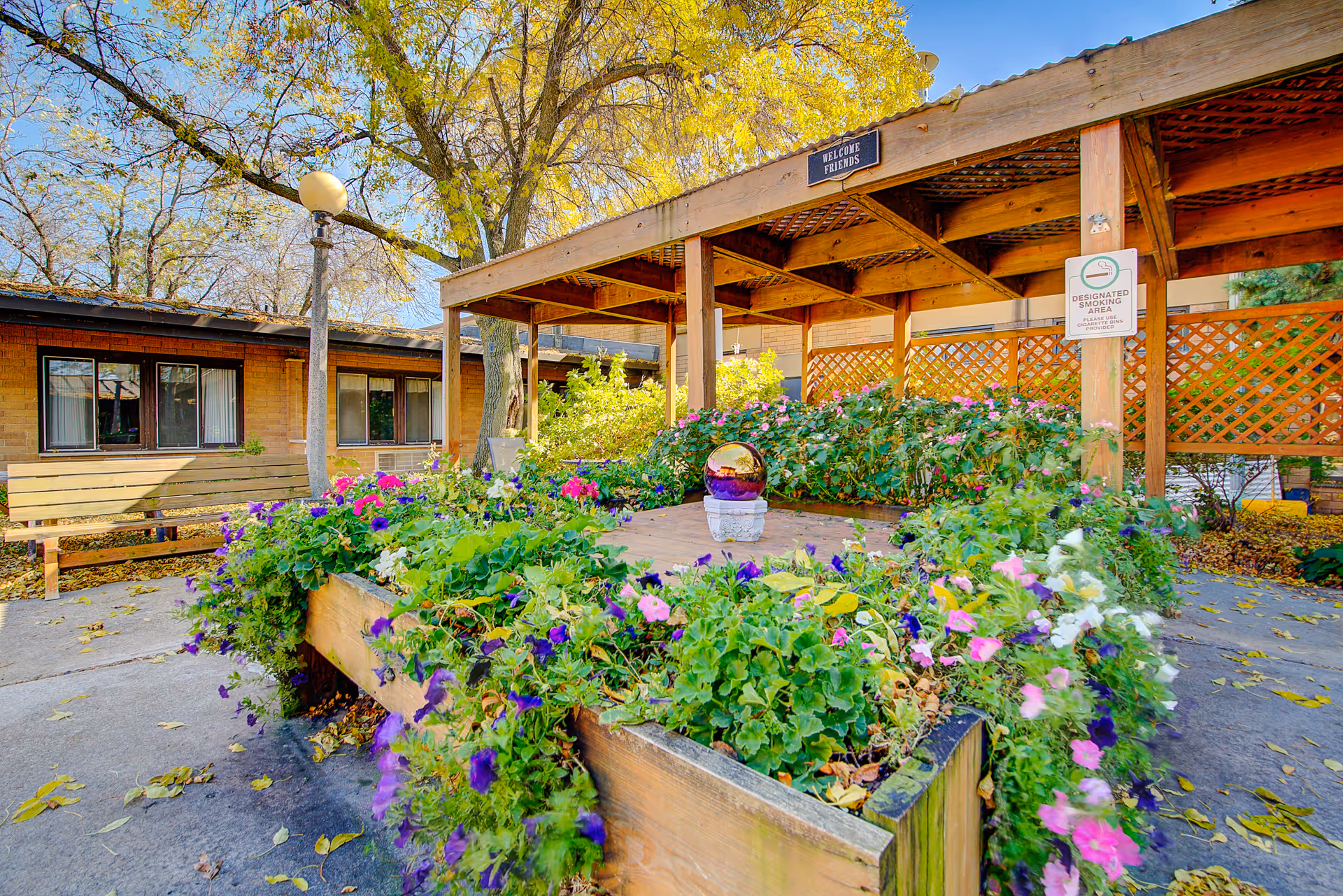 Outdoor garden area at Edenbrook Oshkosh featuring a wooden pergola with lattice sides, a raised flower bed filled with colorful flowers, a bench, and a lamp post. Trees with autumn leaves surround the area under a clear blue sky.