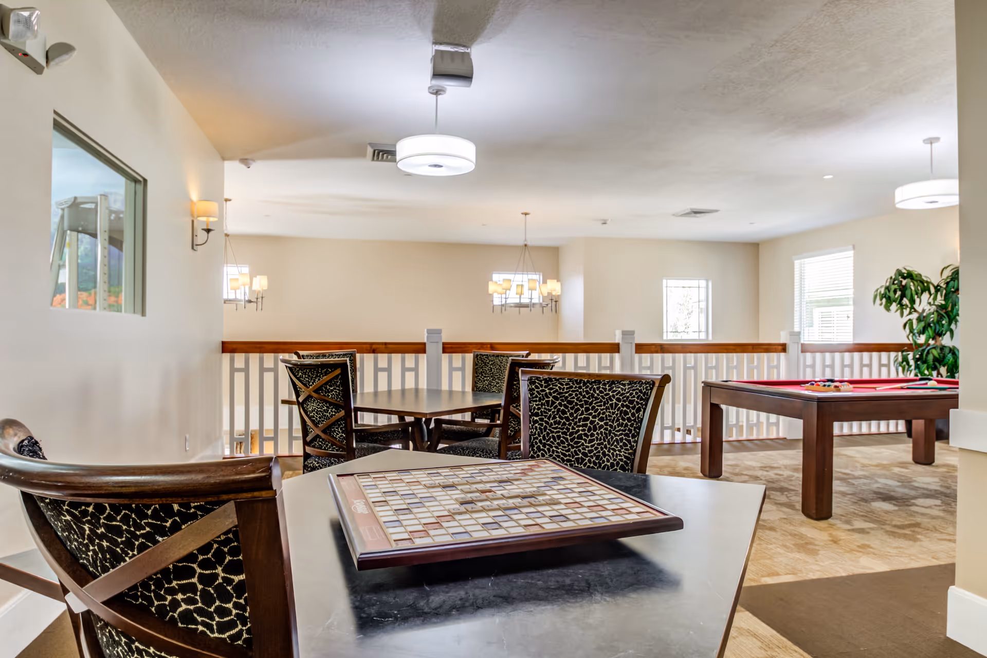 A bright and spacious common area in a senior living facility featuring a table with a Scrabble board, several chairs with patterned upholstery, a pool table with billiard balls, large windows letting in natural light, and modern ceiling lights.