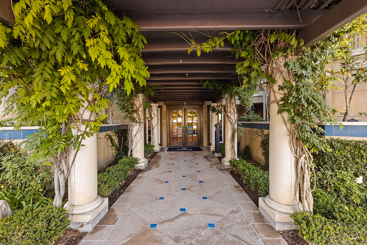 Covered walkway with beige tiled floor and blue square accents, flanked by columns wrapped in green vines and plants, leading to double glass doors at the entrance of a building.