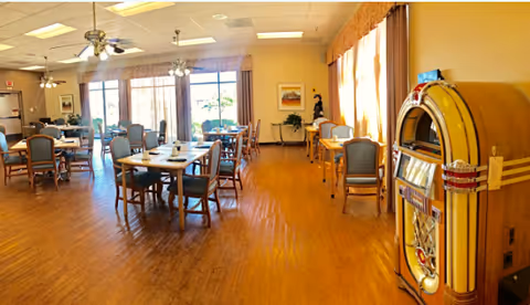 A spacious senior dining room with multiple tables and chairs, large windows letting in light, and a vintage jukebox near the right wall.