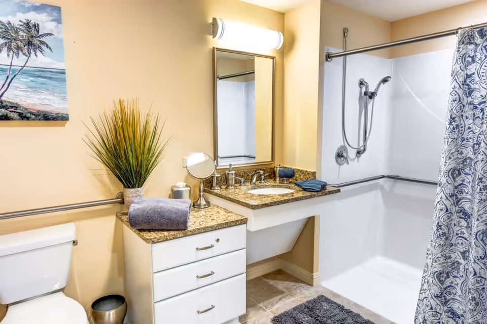 A clean and well-lit bathroom featuring a white toilet, a granite countertop with a sink, a large mirror, and a walk-in shower with a handheld showerhead and a blue and white patterned shower curtain. There is a potted plant and folded towels on the countertop, a small round mirror, and a wall-mounted grab bar near the toilet and in the shower area. The walls are painted light beige, and a painting of a beach scene with palm trees hangs above the toilet.