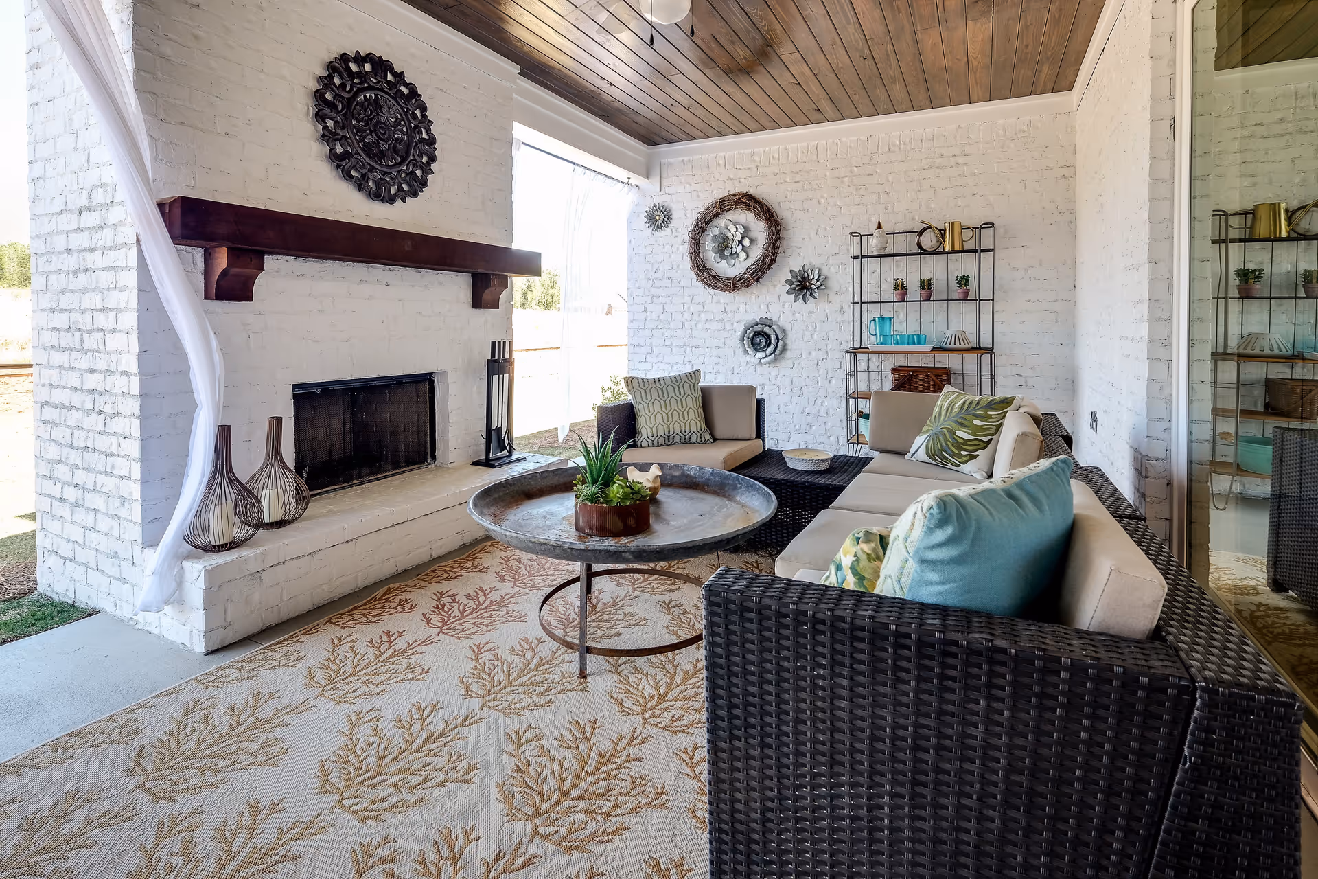 A cozy indoor seating area with wicker furniture including a sofa and chairs with cushions, a round metal coffee table with a plant centerpiece, a white brick fireplace with a wooden mantel, decorative wall hangings, and a patterned rug with coral designs. The space has a wooden ceiling and large windows letting in natural light.