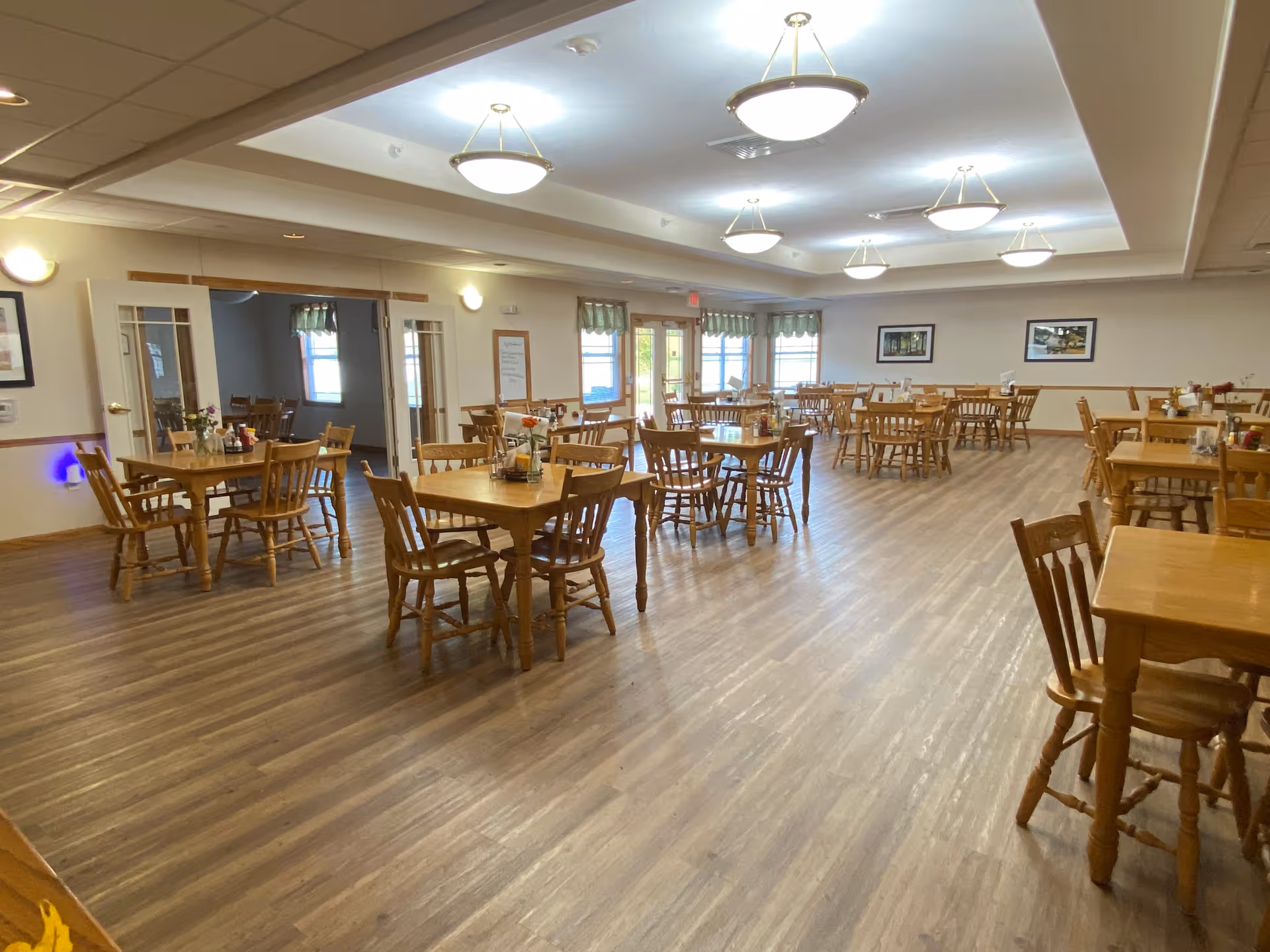 A spacious dining room with multiple wooden tables and chairs arranged neatly on a wood-patterned floor. The room is well-lit with ceiling lights and has large windows with green valances letting in natural light. There are framed pictures on the walls and a whiteboard near the entrance.