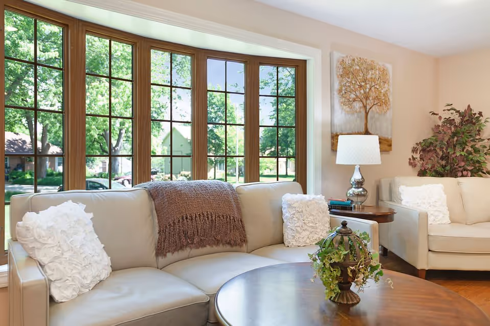 Bright living room with beige sofas, white decorative pillows, a wooden coffee table and large multi-pane windows overlooking a green yard.