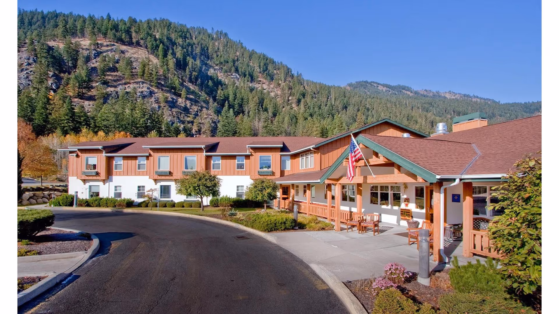 Exterior view of Mountain Meadows Senior Living Campus with a two-story building featuring a brown and white facade, a covered entrance with an American flag, benches, and a curved driveway. The background shows a forested hillside under a clear blue sky.