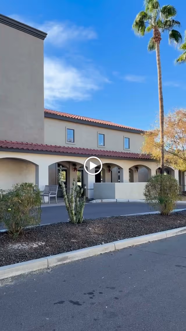 Exterior view of a senior living facility building with beige stucco walls and a red tile roof under a clear blue sky. There are several arched openings along the front with a bench and some desert landscaping including a cactus and bushes. A tall palm tree is visible on the right side.