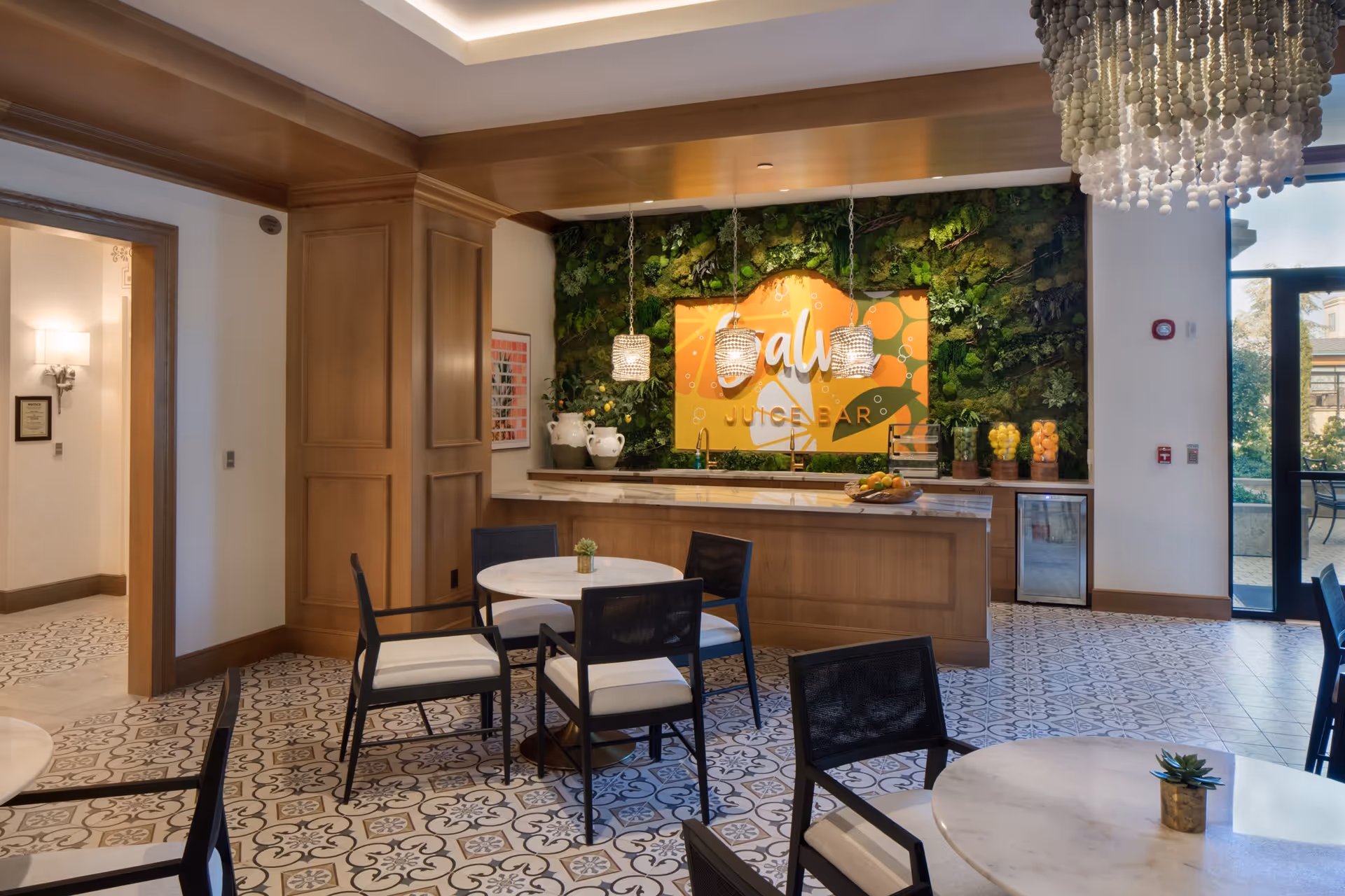 A modern juice bar area inside a senior living facility with a wooden counter, three hanging pendant lights, and a vibrant green plant wall behind it featuring a colorful sign that reads 'Juice Bar'. The room has patterned tile flooring, round tables with black chairs, and a large window letting in natural light.