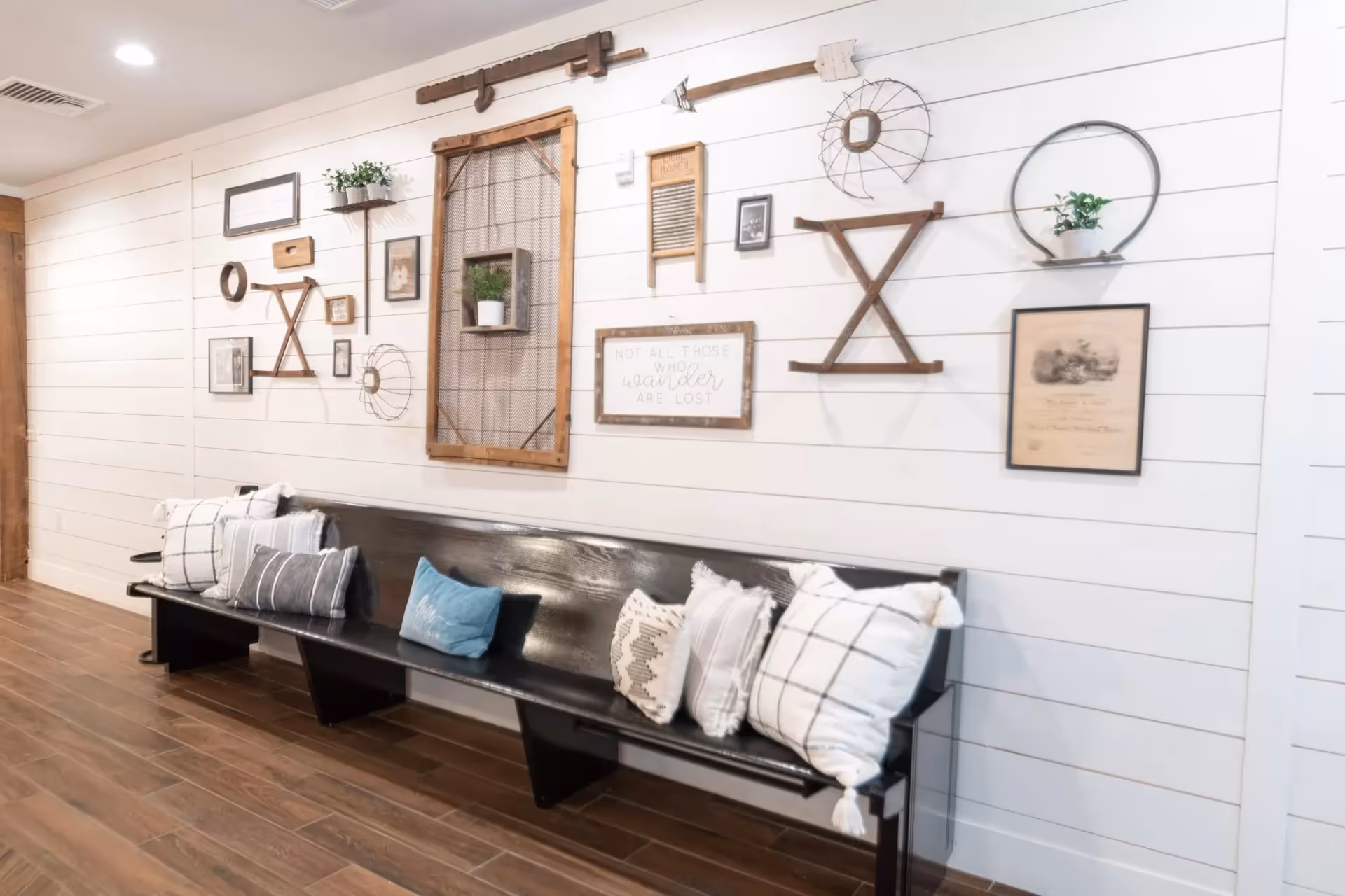 Long black wooden bench with throw pillows against a white shiplap wall decorated with framed art and rustic wall hangings.