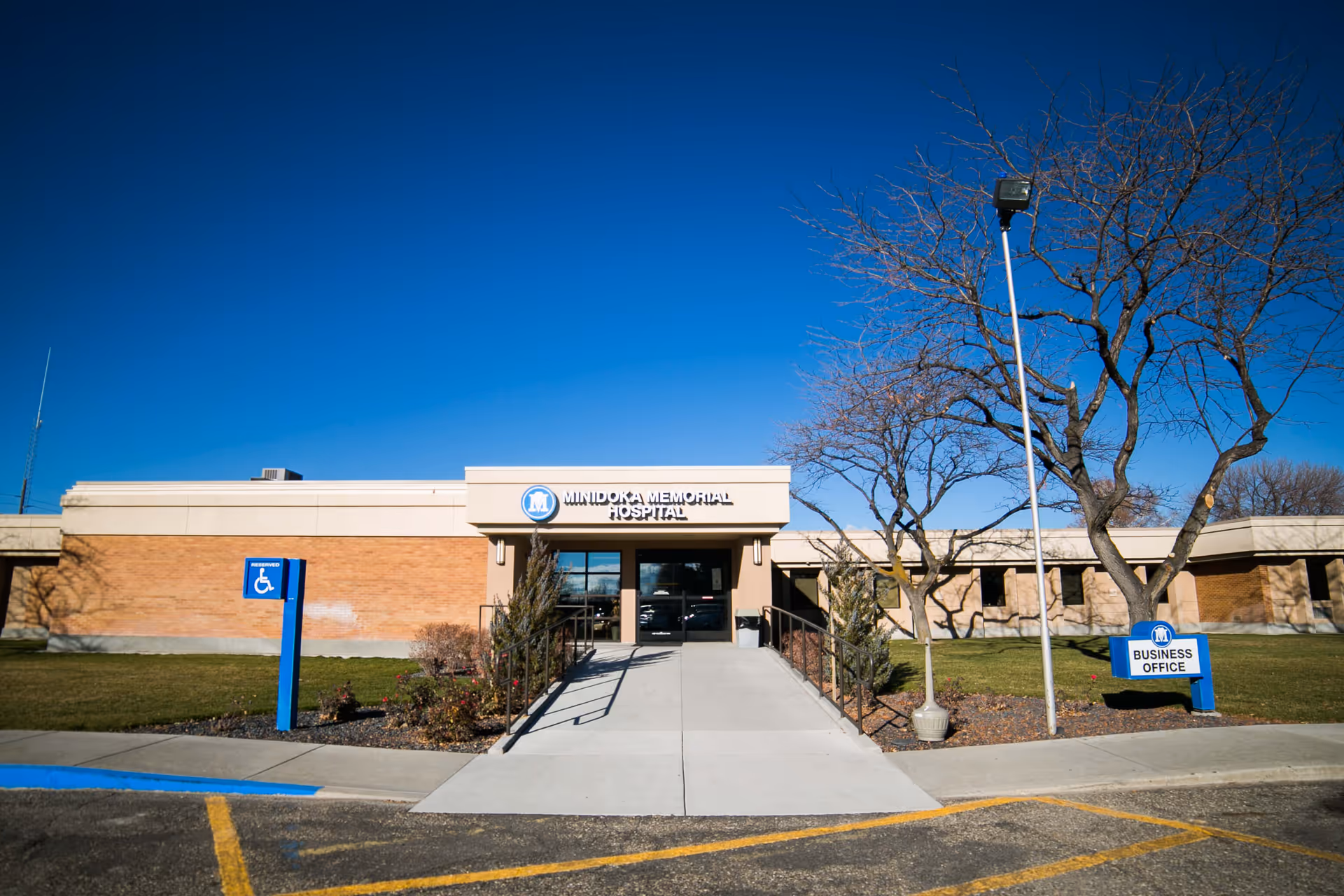 Exterior front view of Minidoka Memorial Hospital building with a clear blue sky. There is a ramp leading to the entrance, a handicapped parking sign on the left, and a business office sign on the right near a leafless tree.