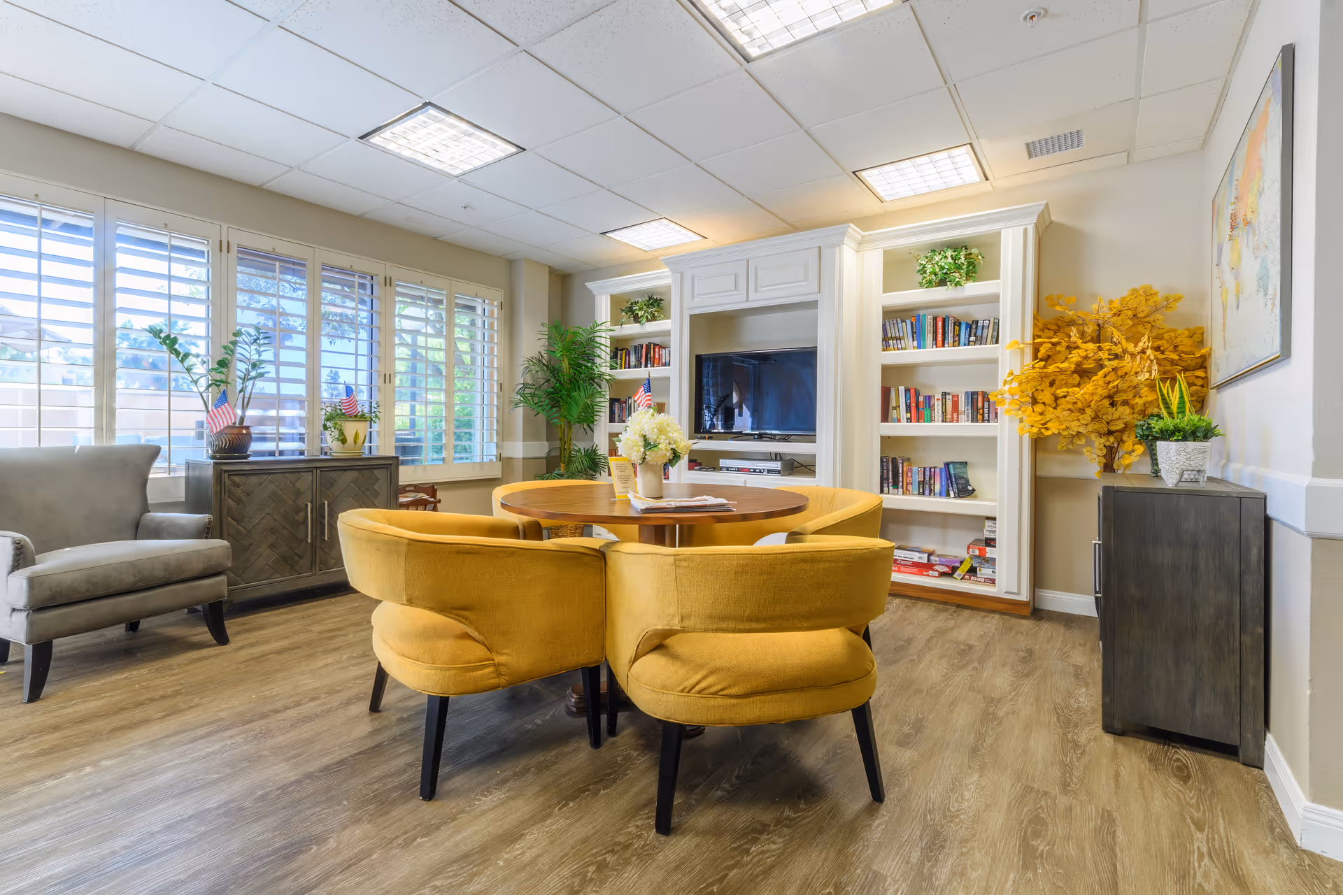 A bright and cozy living room area with a round wooden table surrounded by four mustard yellow upholstered chairs. There is a gray armchair to the left near large windows with white shutters. The back wall features built-in white bookshelves filled with books and a flat-screen TV in the center. Various green plants and a yellow leafy plant add a touch of nature to the room. The floor is wood with a light finish, and the ceiling has recessed lighting panels.
