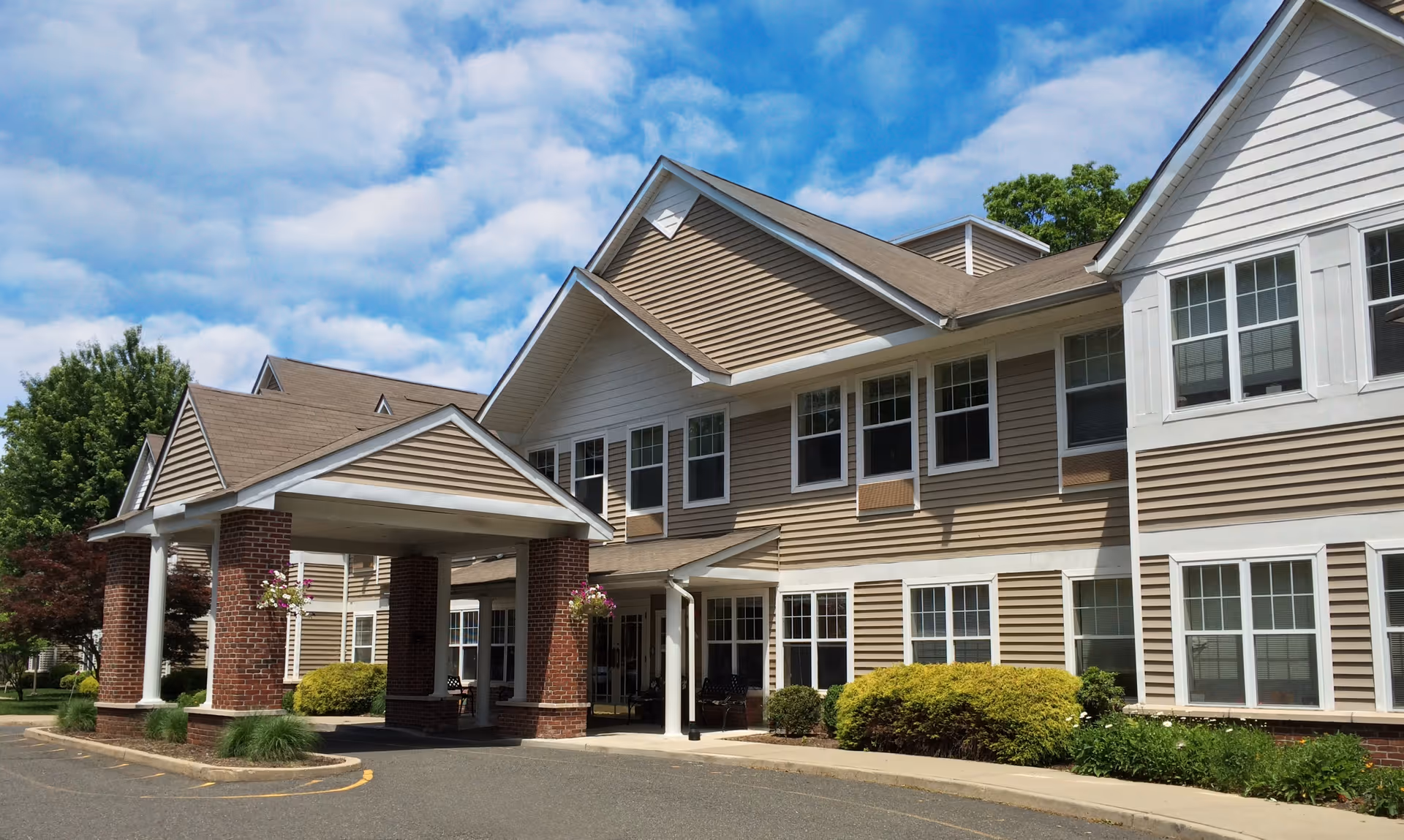 Exterior view of a senior living facility building with beige siding and white trim, featuring a covered entrance supported by brick columns, surrounded by greenery and under a partly cloudy blue sky.