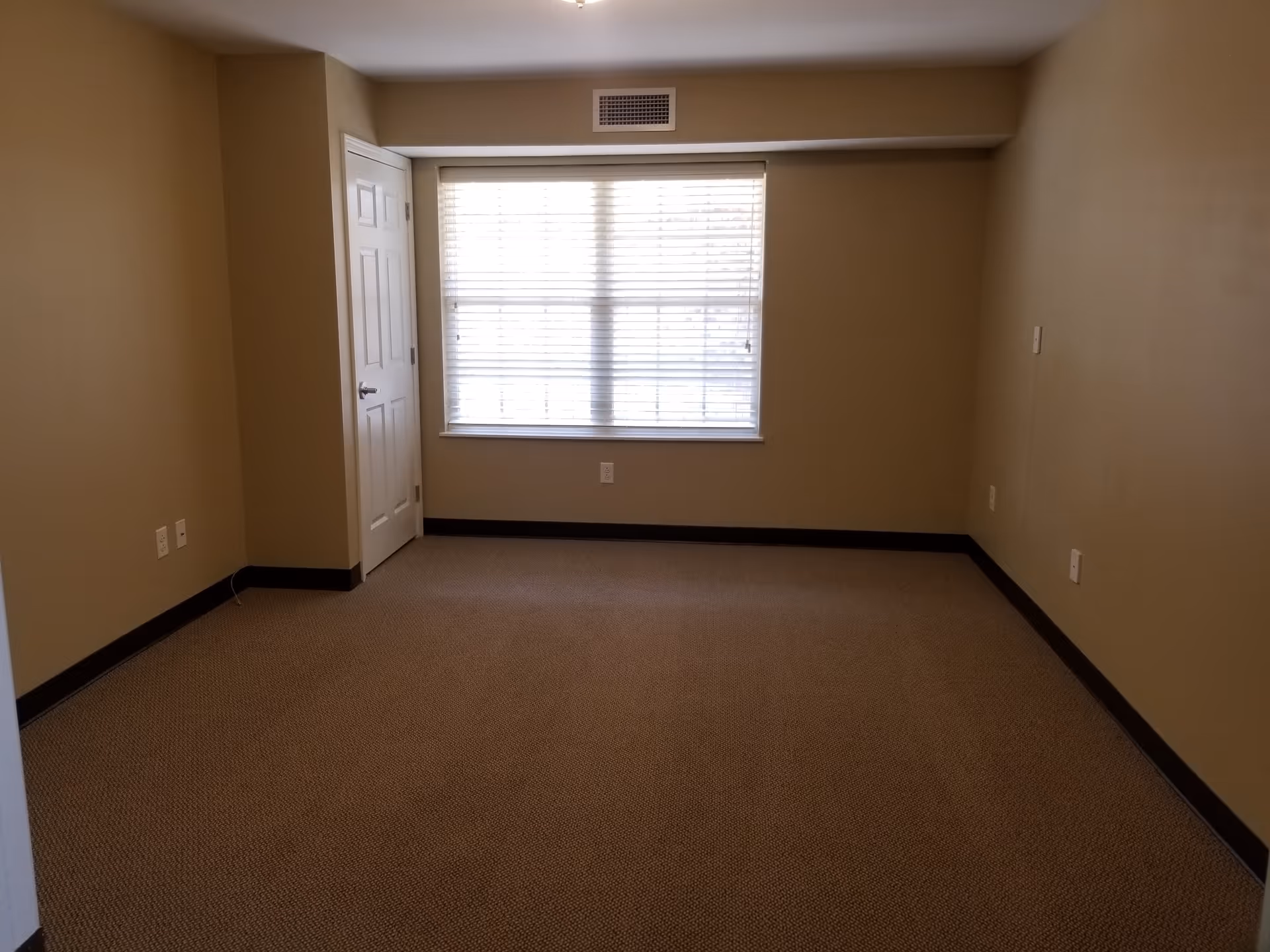 Empty room with beige walls and carpet, a window with white blinds, a white door on the left side, and several electrical outlets along the walls.