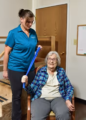 A female caregiver in a blue polo shirt assists an elderly woman seated in a chair by holding a blue exercise band in a room with wooden stairs and a whiteboard on the wall.