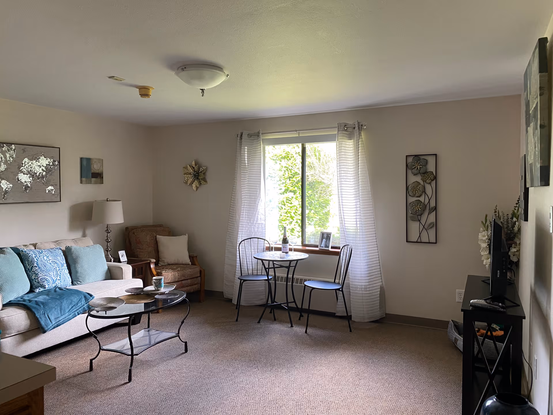 A cozy living room in a retirement community featuring a beige sofa with blue and patterned cushions, a glass coffee table with plates and a cup, a single armchair, a small round table with two chairs near a window with sheer white curtains, and a TV on a stand with decorative flowers beside it. The walls are decorated with framed artwork and metal wall hangings.