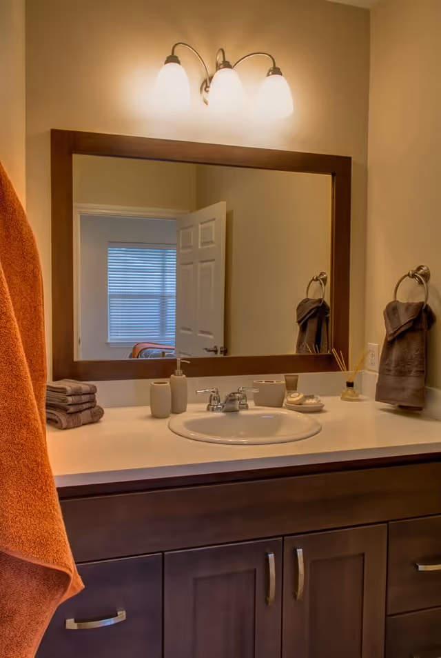 Bathroom sink area with a large rectangular mirror above it, a three-light fixture on the wall, and a countertop holding soap dispensers, a cup, and decorative items. Towels hang on both sides of the sink, and a partially open door and window with blinds are visible in the mirror's reflection.