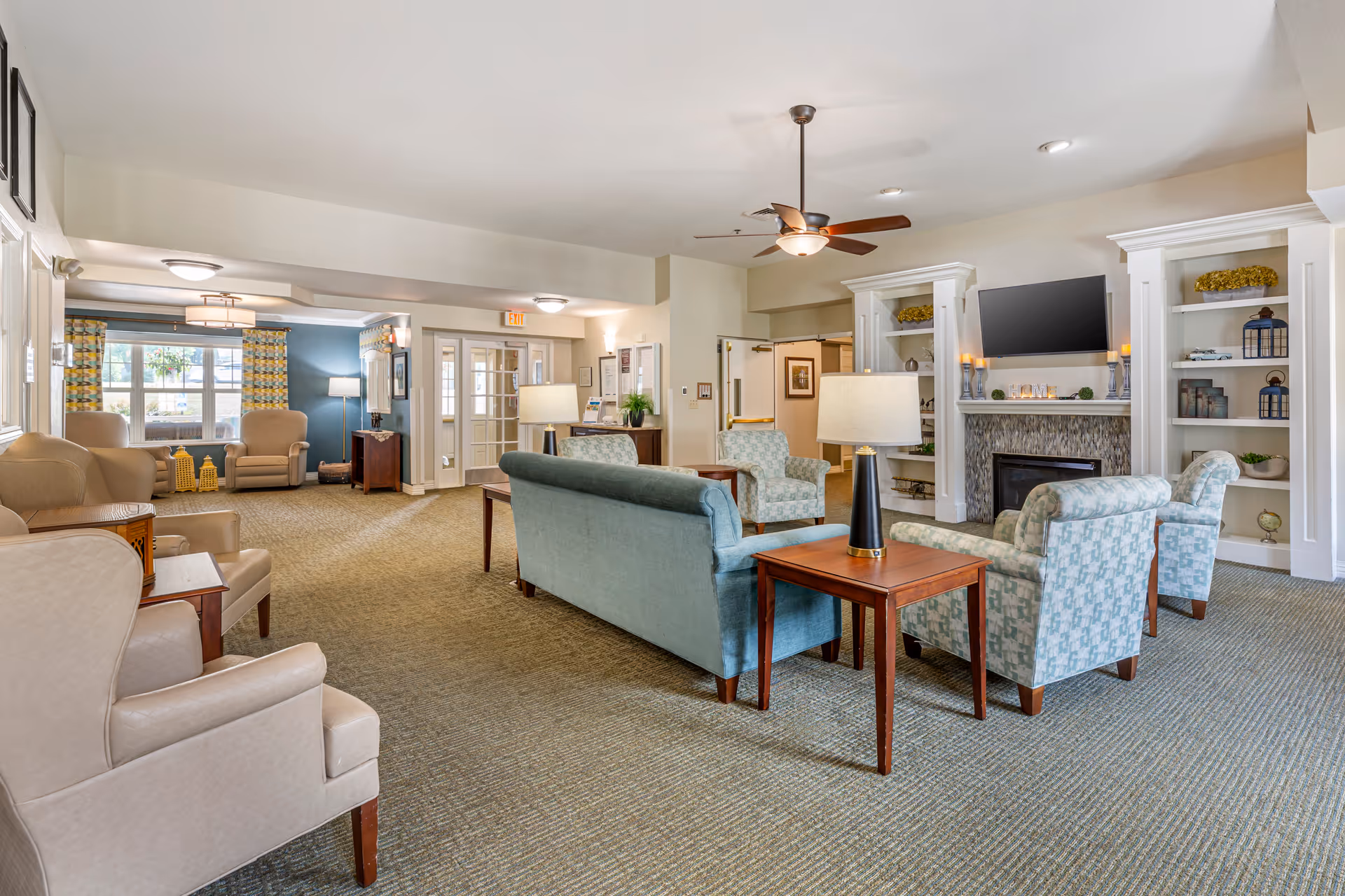 A spacious and well-lit living room area in a senior living facility with multiple armchairs and sofas arranged around a central coffee table. The room features a fireplace with a mounted flat-screen TV above it, built-in shelves with decorative items, and large windows with patterned curtains allowing natural light to enter. There are ceiling lights and lamps providing additional lighting.
