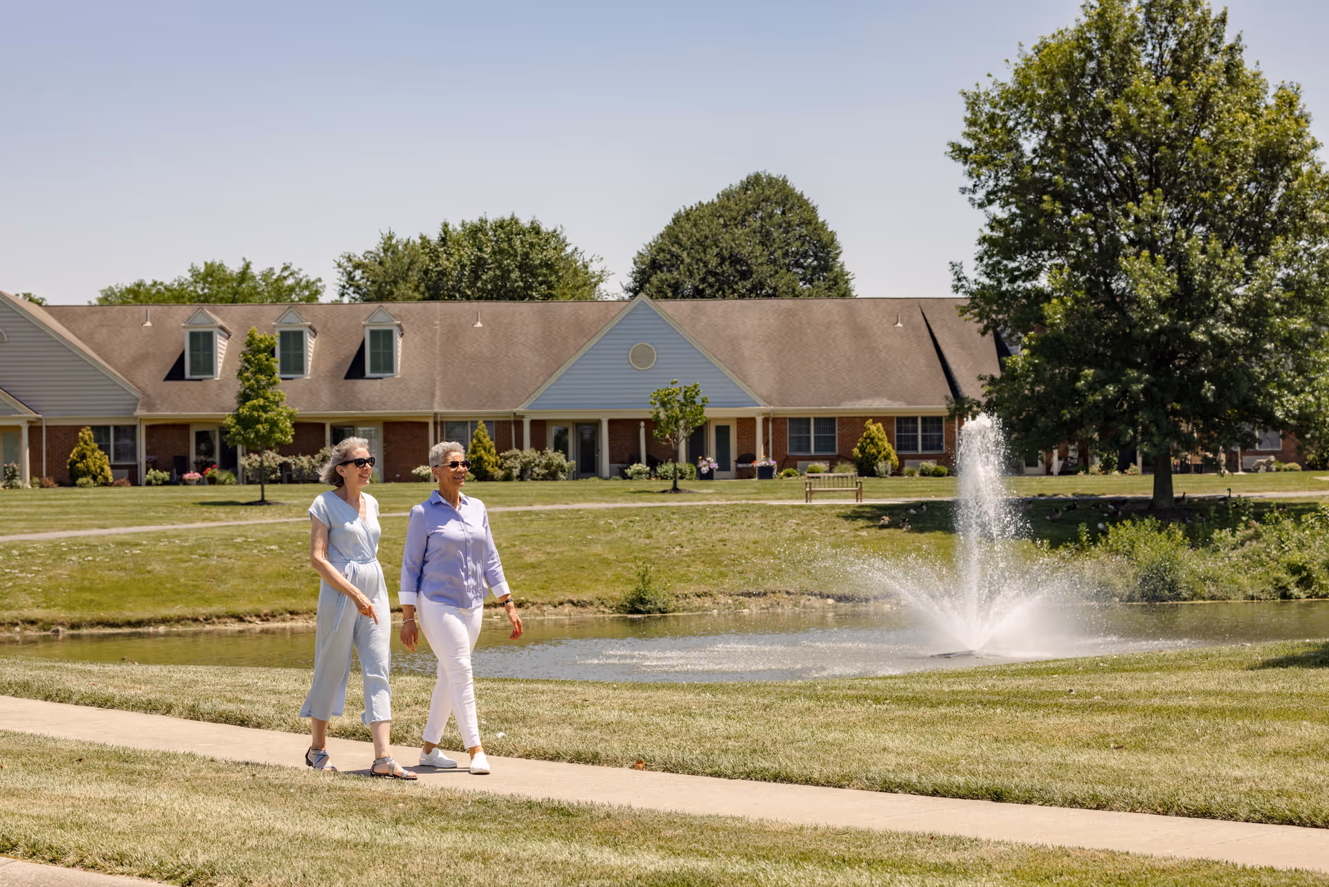 Two elderly women walking and smiling on a paved path next to a pond with a water fountain, with a row of single-story residential buildings and trees in the background under a clear sky.