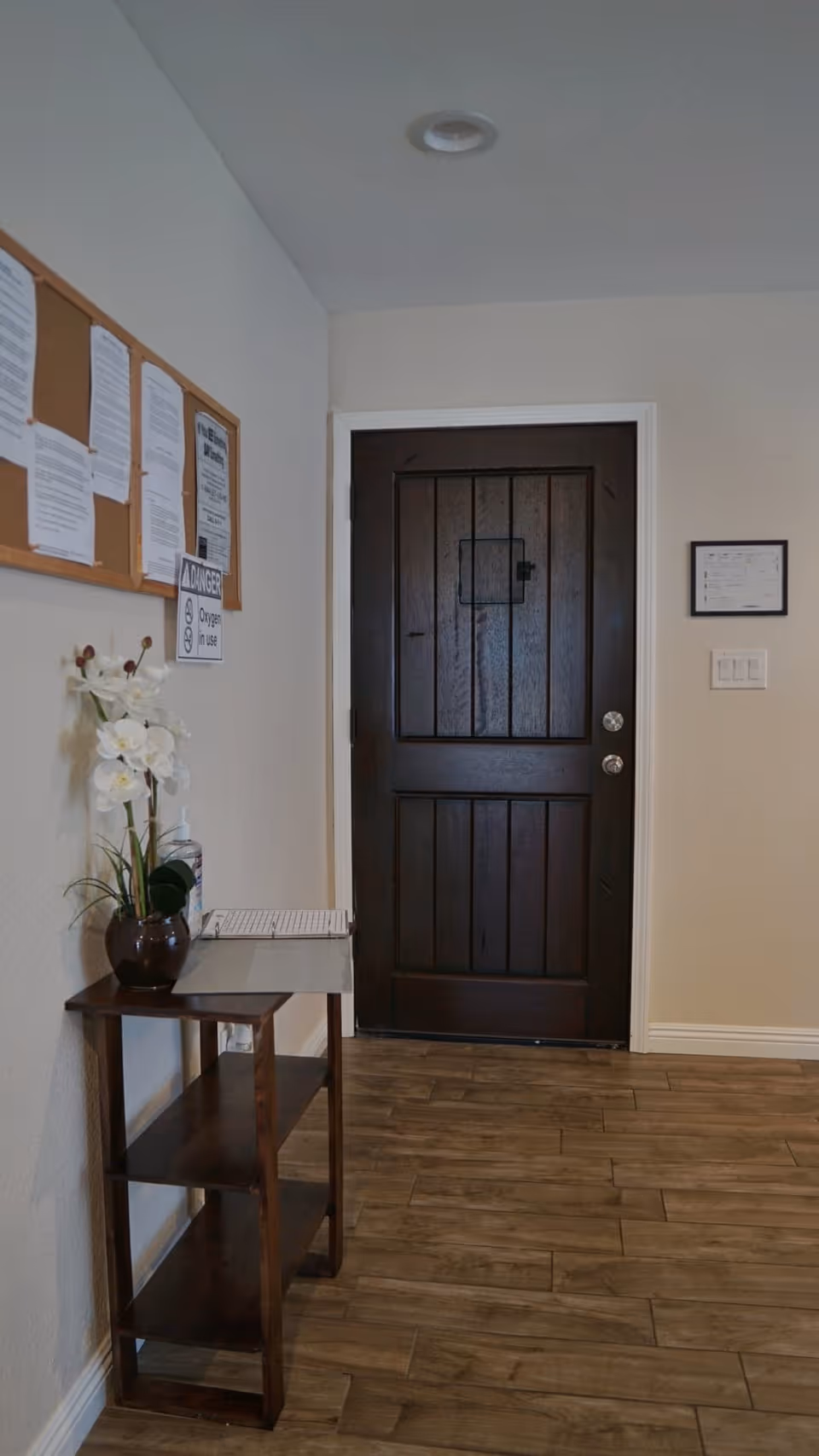 Interior view of a hallway with a dark wooden door at the end. To the left, there is a small wooden table with a potted white orchid plant and a bottle of hand sanitizer. Above the table, a bulletin board with various notices is mounted on the wall. The floor is covered with wood-like tiles.