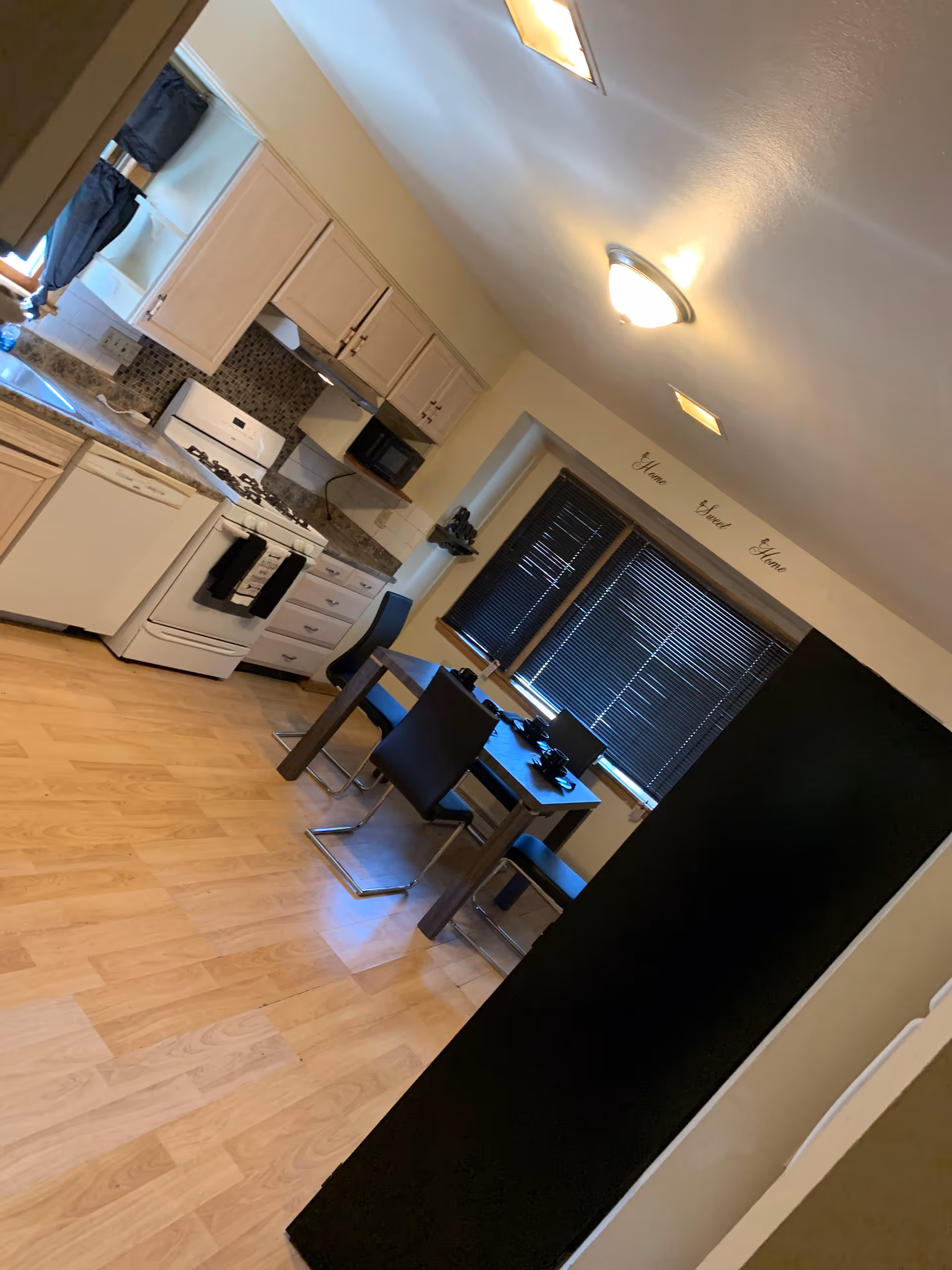 Interior view of a kitchen and dining area with light wood flooring, white cabinets, a white stove, dishwasher, and microwave. A small dining table with four black chairs is positioned near a window with closed black blinds. The wall above the window has the words 'Home Sweet Home' written on it.