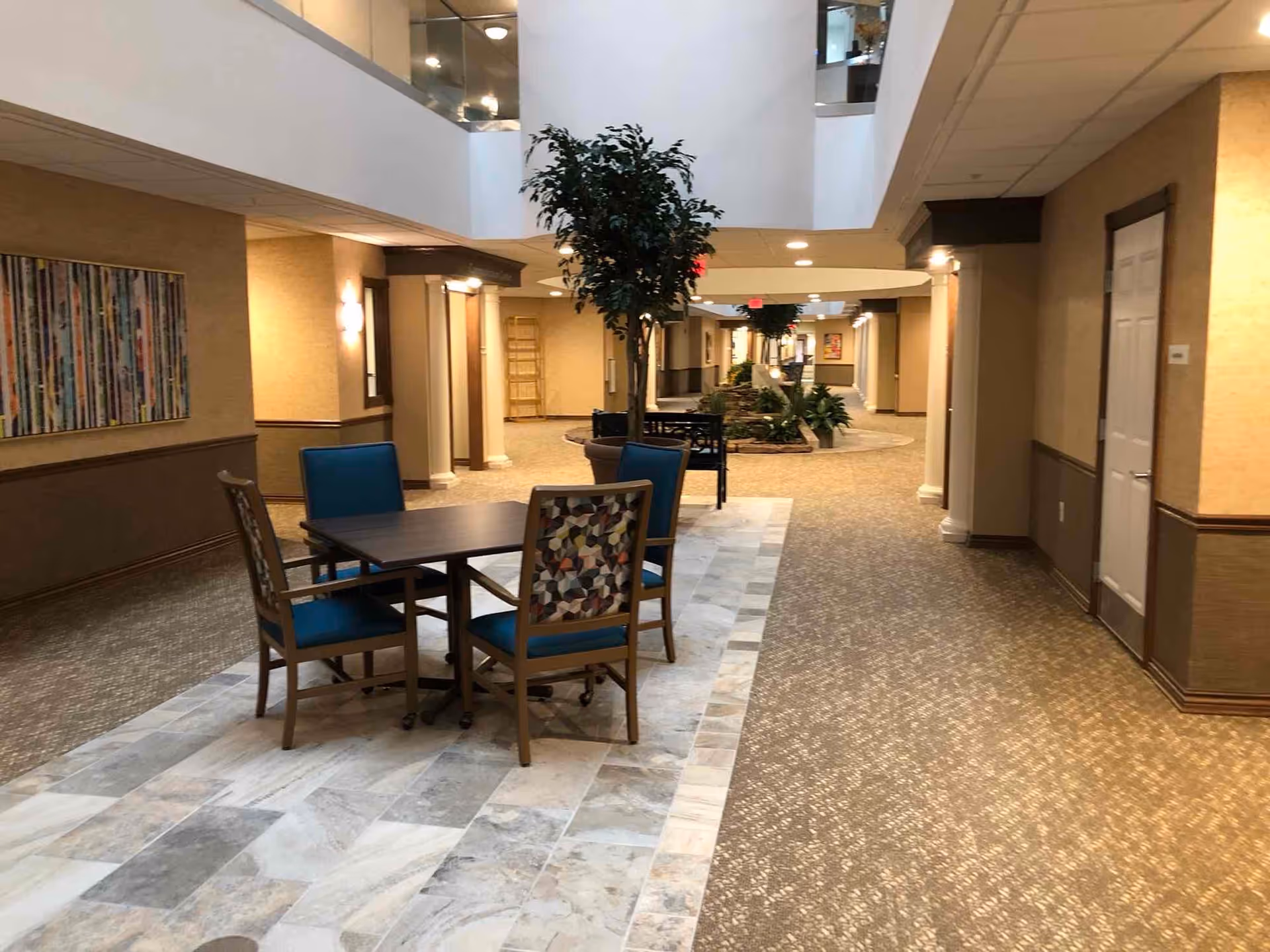 Interior view of a retirement community hallway with a seating area featuring a table and four chairs, two with blue upholstery and two with patterned upholstery. There are potted plants along the hallway, beige walls, carpeted floors, and tiled flooring around the seating area. The hallway is well-lit with ceiling lights and has a second-floor balcony visible above.