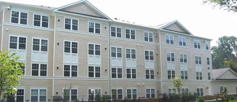 Exterior view of a three-story beige senior living facility building with multiple windows and a small garden area in front.