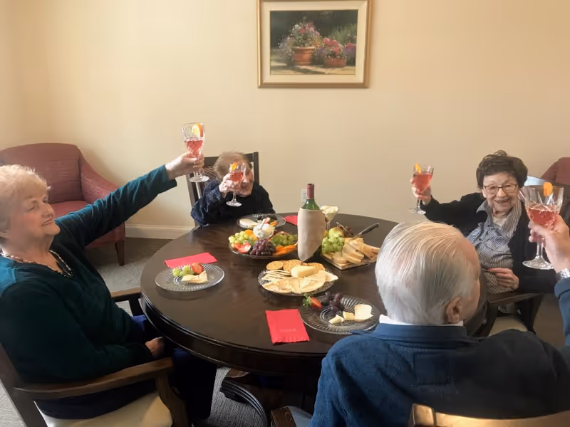 Four elderly residents seated around a round table in a dining room raising glasses in a toast over fruit, cheese, crackers, and a bottle of wine.